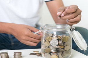 Close-up of hands dropping a coin into a glass jar full of coins on a table.