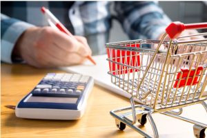 Person writes with a pencil next to a calculator and a miniature shopping cart on a wooden desk.