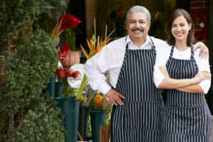 Two smiling shop owners in striped aprons stand outside a flower shop.