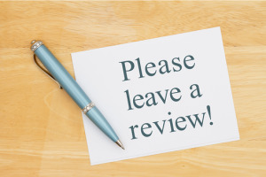 Note card reading 'Please leave a review!' with a blue pen on a wooden desk surface.