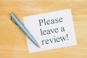 White card on a wooden surface reading 'Please leave a review!' with a blue pen resting diagonally across it.